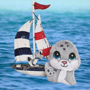 May include: A plush gray seal toy with large blue eyes and a pink nose, next to a miniature sailboat with red and white striped sails and a blue and white striped sail. The background is a blue ocean.