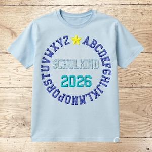 May include: A light blue t-shirt with a circular design. The design includes the word "SCHULKIND" in light blue, the year "2026" in teal, and a ring of blue letters. A yellow star is at the top of the circle.
