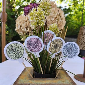 May include: A decorative floral arrangement featuring embroidered felt flower toppers on wooden sticks. The toppers showcase various floral and geometric designs in shades of green, pink, yellow, and blue, set against a white background. The arrangement is in a square, bronze-colored pot.