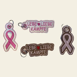 Könnte beinhalten: Vier Schlüsselanhänger mit rosa und lila Schleifen-Designs und dem Text "LEBE LIEBE KÄMPFE" mit einem Herzsymbol.