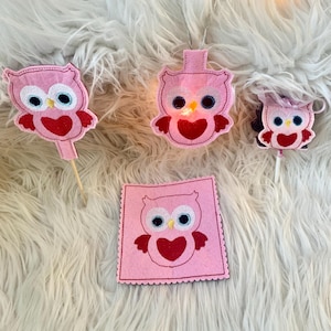 May include: Four pink felt owl ornaments with red hearts. Two owls are on sticks, one is hanging, and one is on a square felt piece.