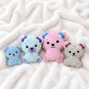 May include: Four plush bear toys in varying sizes and colors. The smallest is light blue with pink accents, followed by a larger light blue bear with purple accents, a pink bear with blue accents, and a gray bear with pink accents.