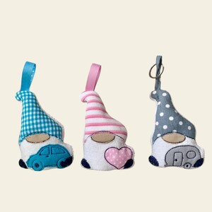 May include: Three handmade gnome ornaments. The gnome on the left has a blue and white gingham hat and a blue car on its belly. The gnome in the middle has a pink and white striped hat and a pink heart on its belly. The gnome on the right has a gray and white polka dot hat and a gray camper on its belly.