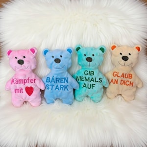 Könnte beinhalten: Vier farbige Plüschteddys mit gesticktem Text. Die Bären sind rosa, blau, türkis und beige, jeder mit einer anderen Botschaft. Der rosa Bär hat ein rotes Herz. Die Bären liegen auf einer weißen, flauschigen Oberfläche.