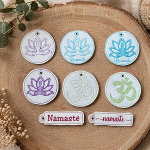 Puede incluir: Parches bordados con temática de yoga con flores de loto y símbolos Om en morado, azul y verde. La palabra "Namaste" también está bordada en un parche rectangular. Los parches están dispuestos sobre una superficie de madera.
