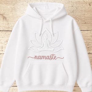 Könnte beinhalten: Weißer Kapuzenpullover mit Fronttasche und Kordelzug. Das Design zeigt eine Lotusblume mit einer sitzenden Figur und dem Wort "namaste" in Schreibschrift. Das Design ist in einem hellrosa Farbton gehalten.