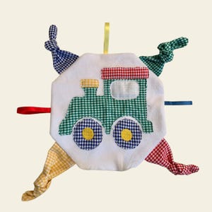 May include: A white, plush, baby toy shaped like a train with a green, red, and yellow checkered pattern. The toy has four knotted fabric ties in blue, red, yellow, and green.