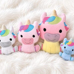 May include: Four plush unicorn toys in varying sizes and colors. The white, pink, yellow, and light blue unicorns feature embroidered details, including black eyes, smiling mouths, and multicolored manes with gold-colored horns on their heads.