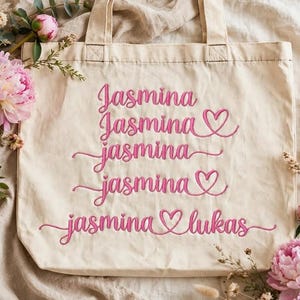 Può includere: Una borsa tote in tela beige con cinque righe di testo ricamato in rosa. Il testo recita Jasmina, Jasmina con un cuore, jasmina, jasmina con un cuore, e jasmina lukas con un cuore che collega i due nomi in un carattere corsivo.