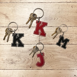 Puede incluir: Llaveros con las letras K, J y M, cada uno unido a un llavero con llaves. La letra K es roja con un contorno blanco, y la letra K es negra con un contorno blanco. La letra J es roja con un contorno blanco, y la letra M es negra con un contorno blanco.
