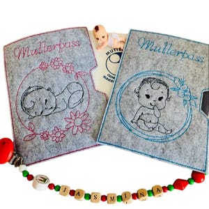 May include: Two gray felt covers with the word "Mutterpass" embroidered on them. Each cover features a different embroidered image of a baby. One baby is sleeping and the other is sitting up. The covers are open to reveal a white card with the word "Mutterpass" and a small doll.