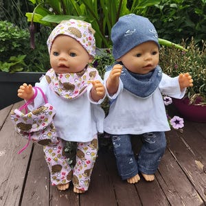 May include: Two baby dolls dressed in handmade outfits. One doll wears a pink hedgehog-themed outfit with matching hat, bib, and purse. The other doll is in a blue denim outfit with a car pattern, a matching hat, and a bib.