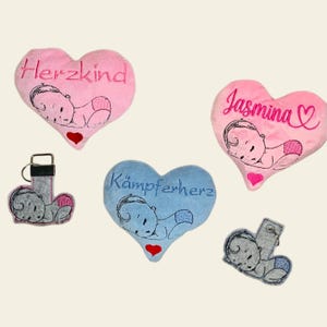 Könnte beinhalten: Drei herzförmige Plüschkissen mit gestickten schlafenden Babys und personalisiertem Text. Die Kissen sind rosa, blau und grau. Der Text auf den Kissen lautet "Herzkind", "Jasmina" und "Kämpferherz".