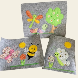 May include: Three gray felt fabric squares with embroidered images of a butterfly, bee, and dragonfly. The squares are decorated with green grass, flowers, and a yellow sun.