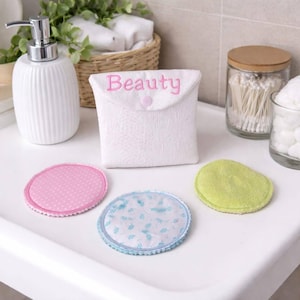 May include: White, textured pouch with the word "Beauty" embroidered in pink, and a pink button closure. Three reusable facial rounds in pink polka dots, blue floral, and green. A white ceramic soap dispenser is on the left.