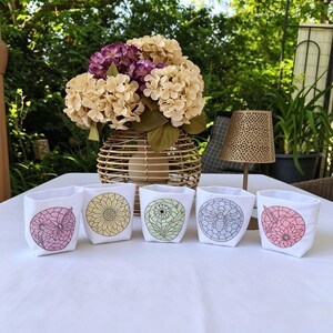 May include: Five white fabric pots with embroidered floral designs, including a sunflower, bee, and abstract patterns. A wicker vase with beige and purple flowers sits behind the pots. The scene is set outdoors on a white tablecloth.