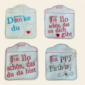 May include: Four white felt pouches with colorful embroidered messages. The messages include "Danke du", "Hallo schön, das es dich gibt", "Hallo schön, das du da bist", and "Happy Birthday".
