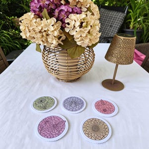 May include: A collection of five decorative coasters with floral and insect designs. The coasters are oval-shaped with white scalloped edges. A woven basket with flowers and a gold-colored lamp are also on the white tablecloth.