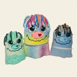 May include: Three plush toys with embroidered faces. The first toy is green with a pink nose and green hair. The second toy is blue with a pink nose and rainbow hair. The third toy is blue with a blue and white checked nose and brown hair.