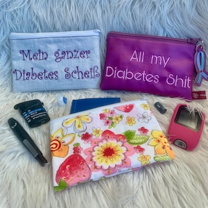 May include: Two zippered pouches, one purple and one white, with the text "All my Diabetes Shit" and "Mein ganzer Diabetes Scheiß" embroidered on them. A third pouch with a floral pattern is also visible, along with a blood glucose meter and other diabetes supplies.