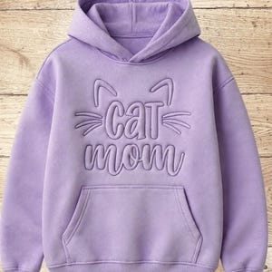 May include: A lavender hoodie with a hood and a front pocket. The design features the words "Cat Mom" in a stylized font, with cat ears and whiskers above the word "Cat".