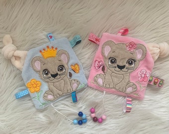 May include: Two plush baby toys with embroidered lion faces. The blue toy has a crown and a butterfly, while the pink toy has flowers and a butterfly. Both toys have a loop for attaching a pacifier or other items.