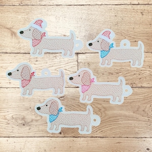 May include: Five dachshund dog ornaments with light brown bodies, each with a different colored bandana and some wearing Santa hats. The ornaments are outlined in light blue and have a small loop for hanging.