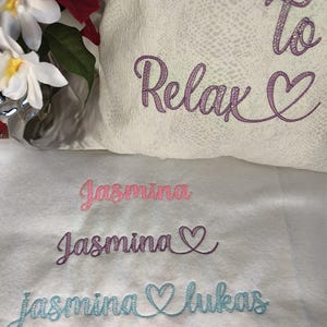 May include: A white pillow with the words "Relax to" embroidered in purple glitter thread, with a heart symbol. Below are three embroidered names: "Jasmina" in pink, "Jasmina" with a heart, and "Jasmina Lukas" in blue.