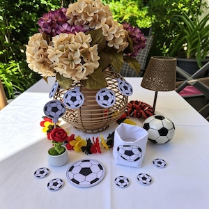 May include: A decorative arrangement featuring a soccer theme. A woven vase holds beige and purple flowers, adorned with soccer ball cutouts. A soccer ball, a small white container with a soccer ball design, and other soccer-themed decorations are on a white tablecloth.