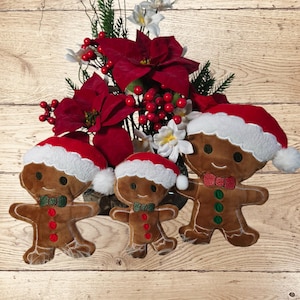 May include: Three plush gingerbread men wearing Santa hats and bow ties, with a floral arrangement of red poinsettias, berries, and white flowers in the background. The gingerbread men have embroidered details.