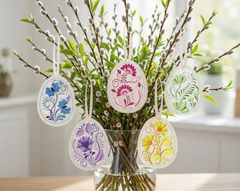 May include: A bouquet of pussy willow branches and green leaves in a glass vase, with five hanging Easter egg ornaments. The egg ornaments are white with colorful floral designs in blue, pink, purple, yellow, and green.