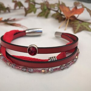 Könnte beinhalten: Ein rotes Lederarmband mit einem roten Kristall-Akzent und silbernen Perlen. Das Armband hat einen Magnetverschluss.
