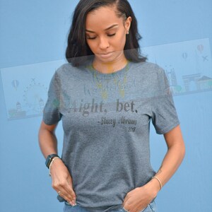 May include: A woman wearing a gray t-shirt with the text "Alright, bet. -Stacey Abrams 2018" and blue jeans. She is standing in front of a light blue wall.