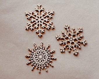 Op de afbeelding: Drie houten sneeuwvlokken in verschillende ontwerpen liggen op een neutrale bruine achtergrond. De sneeuwvlokken hebben ingewikkelde, gedetailleerde patronen. Deze decoratieve elementen zijn ideaal voor wintervakantieknutselwerkjes en decoraties.