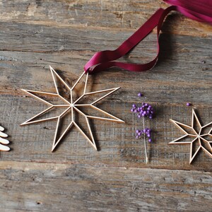 Op de afbeelding: Houten sterornamenten met een gouden afwerking, opgehangen aan een bordeauxrood lint. De sterren zijn geplaatst op een rustiek houten oppervlak, samen met paarse bessen. Een feestelijk, decoratief thema.