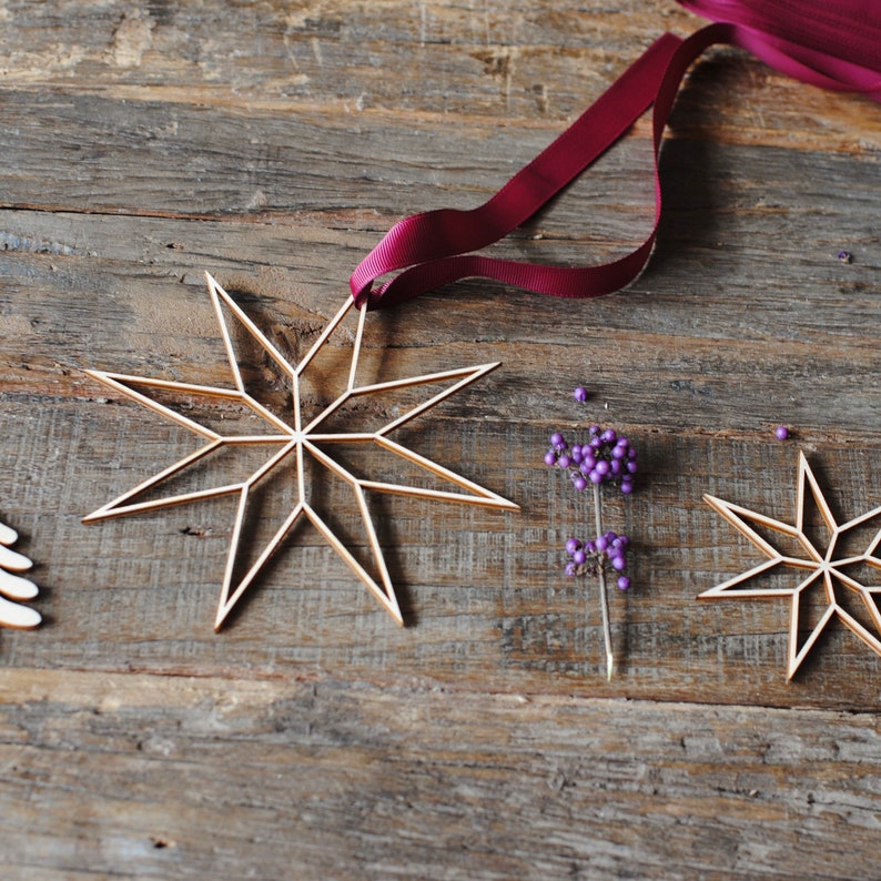 Op de afbeelding: Houten sterornamenten met een gouden afwerking, opgehangen aan een bordeauxrood lint. De sterren zijn geplaatst op een rustiek houten oppervlak, samen met paarse bessen. Een feestelijk, decoratief thema.