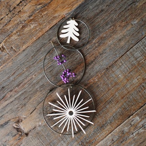 Op de afbeelding: Drie decoratieve metalen ringen verticaal verbonden. De bovenste ring heeft een witte boomuitsparing, de middelste ring heeft paarse bessen en de onderste ring heeft een wit sterrenburst-ontwerp. De ringen staan tegen een rustieke houten achtergrond.