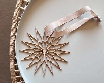 Op de afbeelding: Een lichtbruin, lasergesneden houten sterornament met een geometrisch ontwerp met meerdere punten. De ster is bevestigd aan een glad, lichtbeige lint dat in een eenvoudige lus is geknoopt, wat zorgt voor een strakke en neutrale uitstraling.