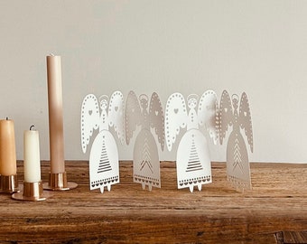Op de afbeelding: Een decoratieve opstelling van witte papieren engelenfiguren met hartvormige vleugels en gedetailleerde jurken. Verschillende kaarsen in verschillende tinten beige en bruin staan op een houten oppervlak, wat een warme, uitnodigende sfeer creëert. De engelen zijn een feestelijke, seizoensgebonden decoratie.