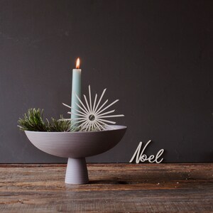 Op de afbeelding: Een decoratief arrangement met een brandende lichtblauwe kaars, een sterornament en dennentakken in een grijze voetbakschaal. Het woord "Noel" staat in witte letters op een houten oppervlak.