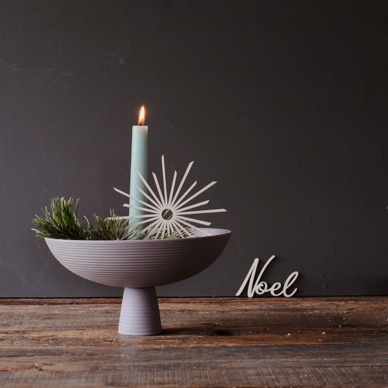 Op de afbeelding: Een decoratief arrangement met een brandende lichtblauwe kaars, een sterornament en dennentakken in een grijze voetbakschaal. Het woord "Noel" staat in witte letters op een houten oppervlak.