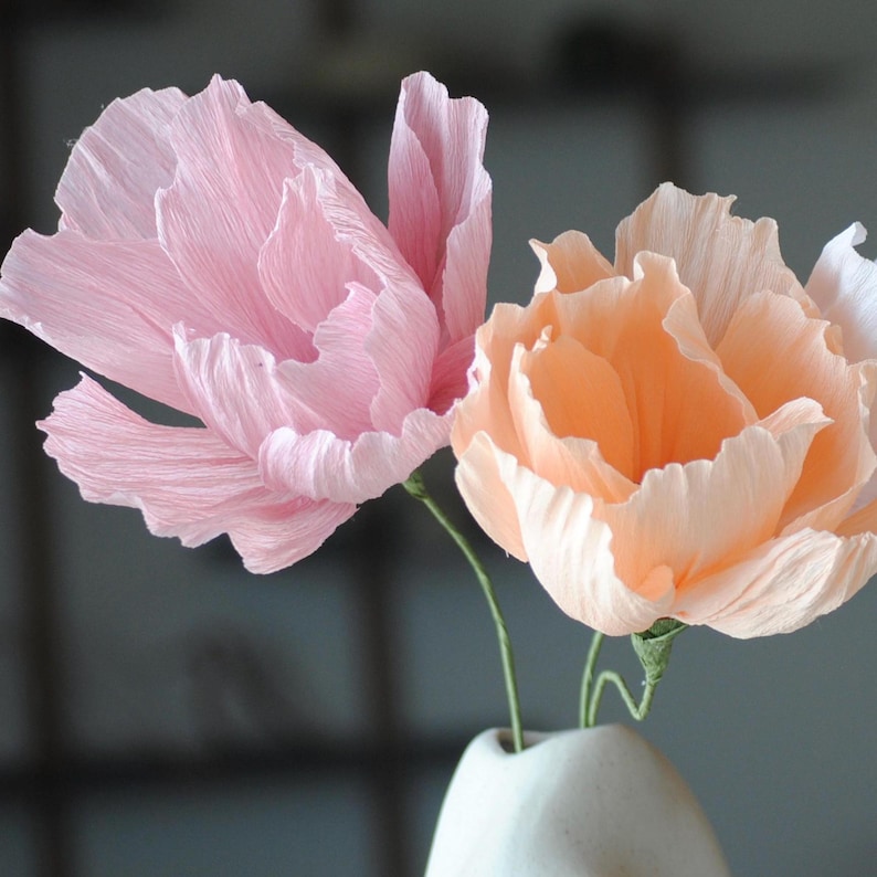 Puede incluir: Dos flores de papel en un jarr&oacute;n blanco. Una flor es rosa claro, la otra melocot&oacute;n. Los p&eacute;talos son texturizados y en capas, creando un aspecto realista. Los tallos son verdes y el jarr&oacute;n es de cer&aacute;mica.