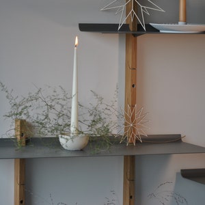 Op de afbeelding: Een modern, minimalistisch rek met grijze metalen planken en houten steunen. Een brandende witte kaars staat in een decoratieve houder, met groen. Decoratieve stervormige ornamenten en een kleine huisvormige decoratie zijn ook aanwezig.
