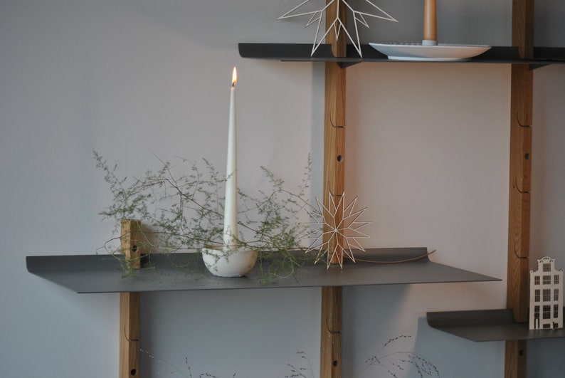 Op de afbeelding: Een modern, minimalistisch rek met grijze metalen planken en houten steunen. Een brandende witte kaars staat in een decoratieve houder, met groen. Decoratieve stervormige ornamenten en een kleine huisvormige decoratie zijn ook aanwezig.