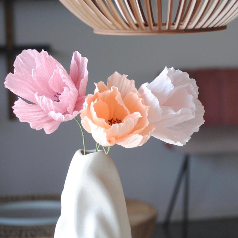 Puede incluir: Un jarr&oacute;n de cer&aacute;mica blanco contiene tres flores de papel en tonos rosa y melocot&oacute;n. Las flores tienen p&eacute;talos delicados y rizados, creando una presentaci&oacute;n suave y elegante. En el fondo se aprecia una pantalla de l&aacute;mpara tejida.