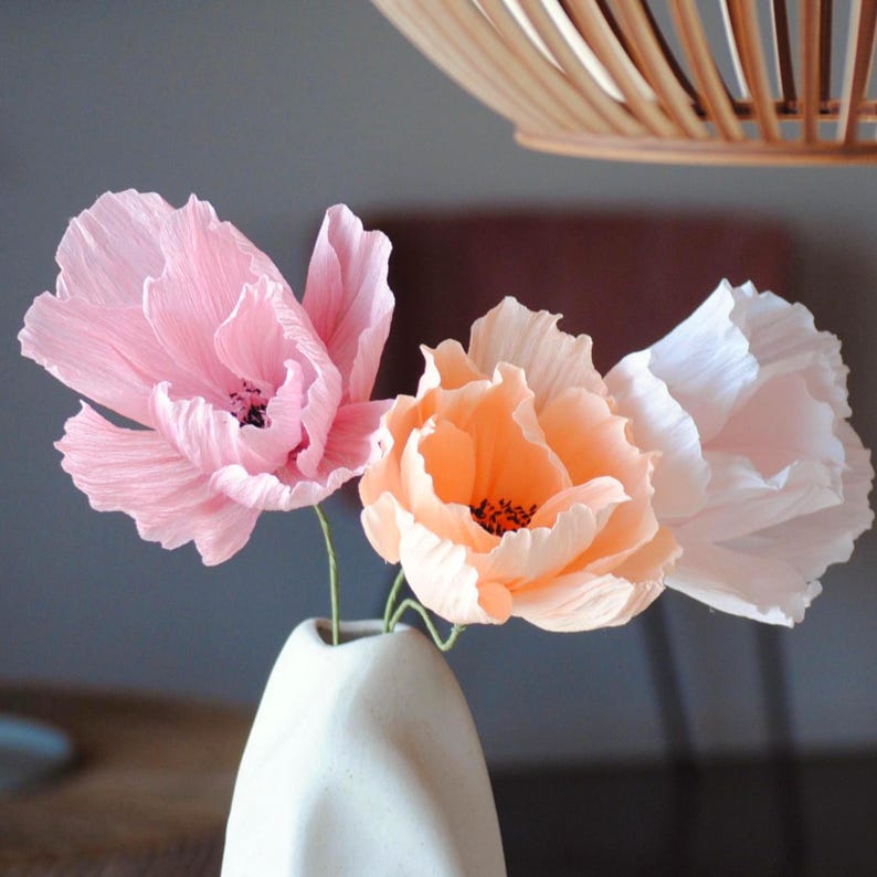Puede incluir: Un jarr&oacute;n de cer&aacute;mica blanco contiene tres flores de papel crep&eacute;. Las flores son en tonos rosa, melocot&oacute;n y blanco, con centros oscuros. Las flores est&aacute;n dispuestas en una presentaci&oacute;n decorativa.
