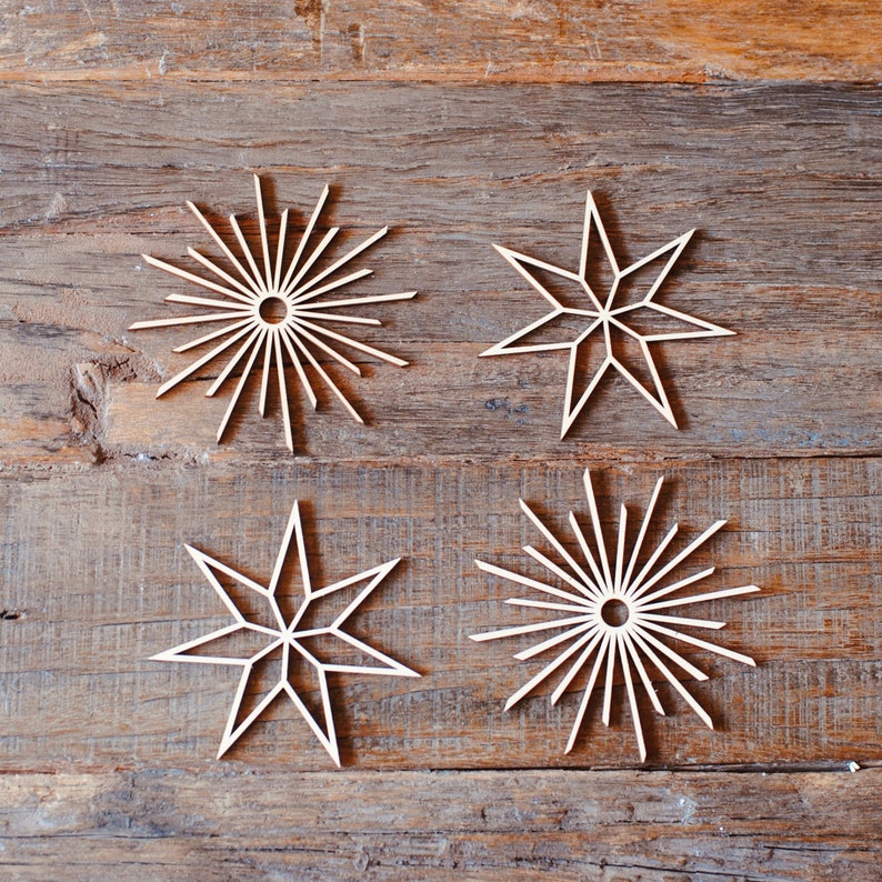 Op de afbeelding: Vier houten sterornamenten in verschillende ontwerpen, gerangschikt op een rustiek houten oppervlak. Twee sterren hebben een sunburst-ontwerp, terwijl de andere twee een meer traditionele stervorm hebben. De natuurlijke houtkleur zorgt voor een warme, neutrale toon.
