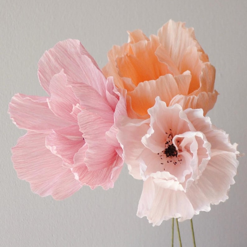 Puede incluir: Tres flores de papel crep&eacute; en tonos rosa y melocot&oacute;n. Las flores tienen p&eacute;talos delicados y rizados y centros oscuros. Los tallos son finos y verdes. Las flores est&aacute;n dispuestas sobre un fondo gris claro.