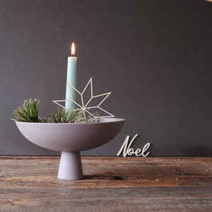Op de afbeelding: Een feestelijk middelpunt met een lichtblauwe kaars, een witte sterdecoratie en dennentakken in een grijze kom. De kom staat op een voetstuk, met het woord "Noel" in cursief.