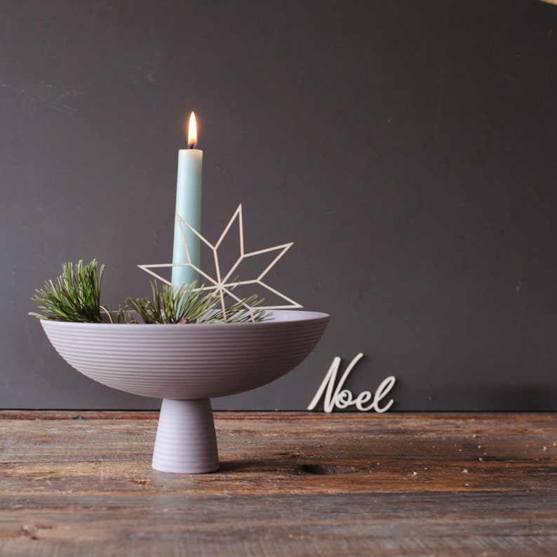 Op de afbeelding: Een feestelijk middelpunt met een lichtblauwe kaars, een witte sterdecoratie en dennentakken in een grijze kom. De kom staat op een voetstuk, met het woord "Noel" in cursief.